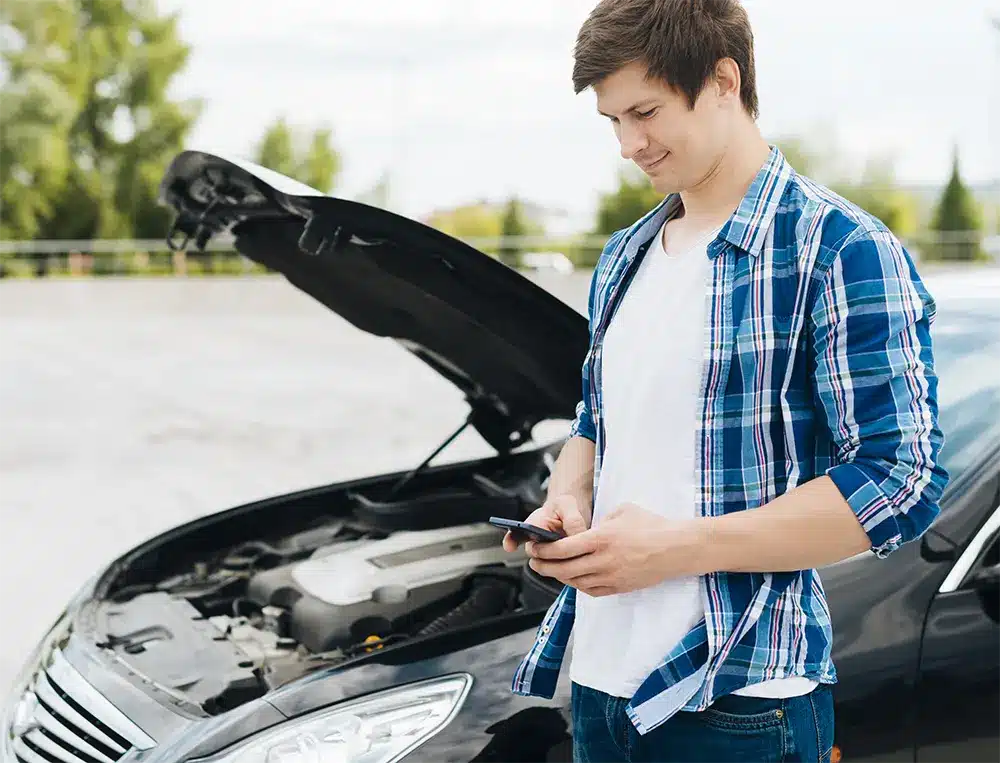 Man in a blue checkered shirt standing by a car with its hood open, looking at his phone, possibly referencing car troubleshooting or roadside assistance.