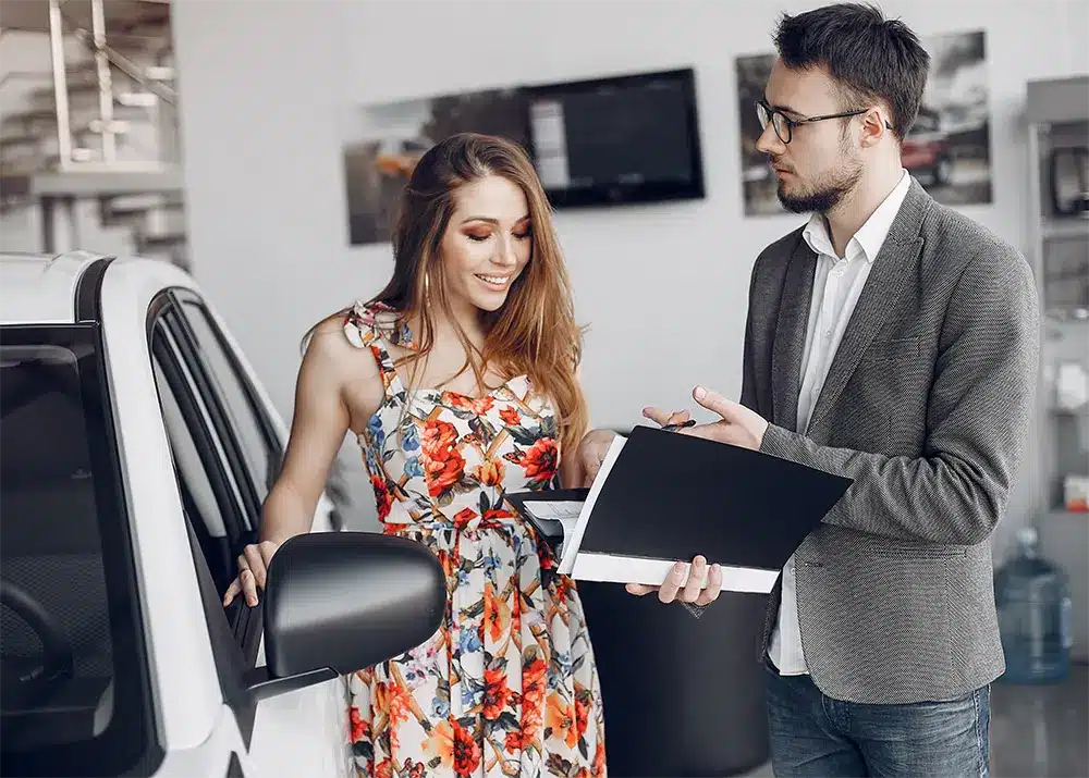 A woman in a floral dress reviewing documents with a man in a gray blazer, standing next to a white car in a professional setting, suggesting a car purchase or consultation.