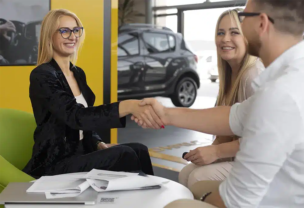 blog-strip A professional woman shaking hands with a couple at a car dealership, with vehicles in the background.