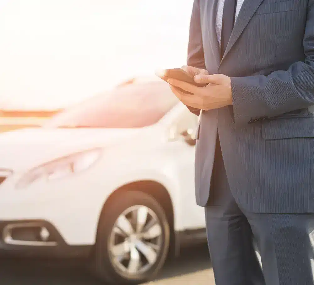 Man in a suit holding a phone in front of a car, symbolizing professional support for Lemon Law cases.