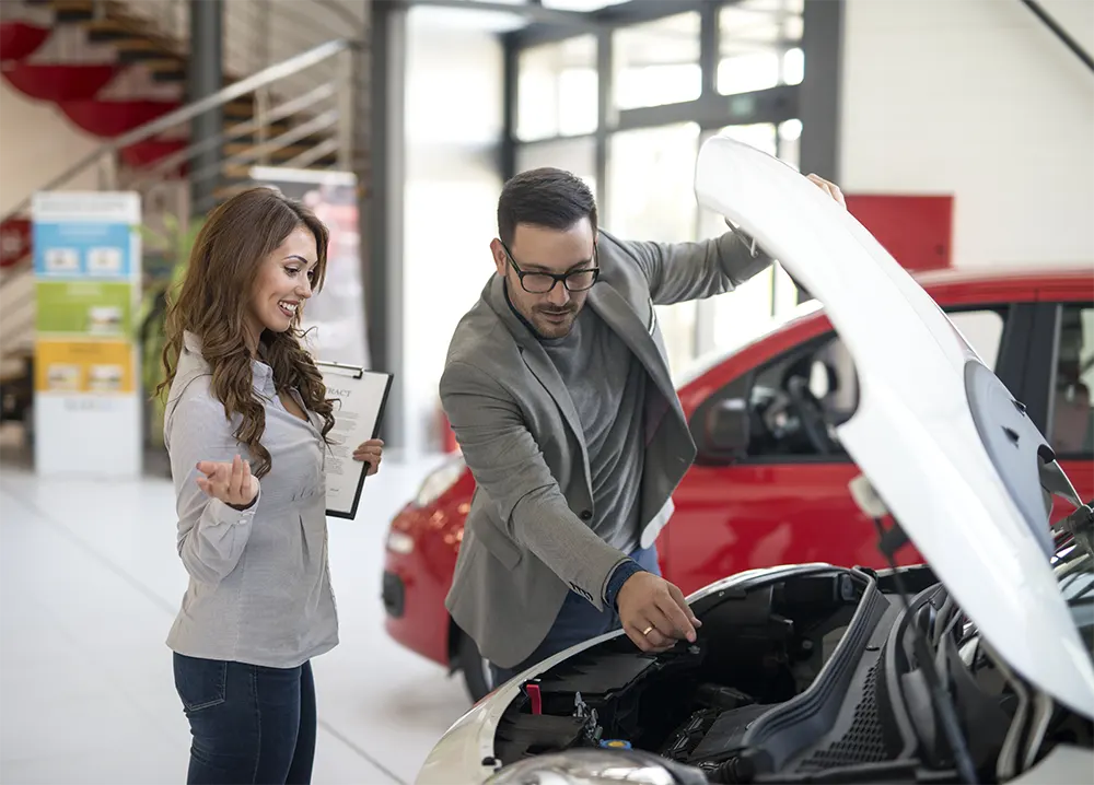 A man in a gray blazer and glasses inspecting the engine of a car while a woman holds a clipboard and discusses details.